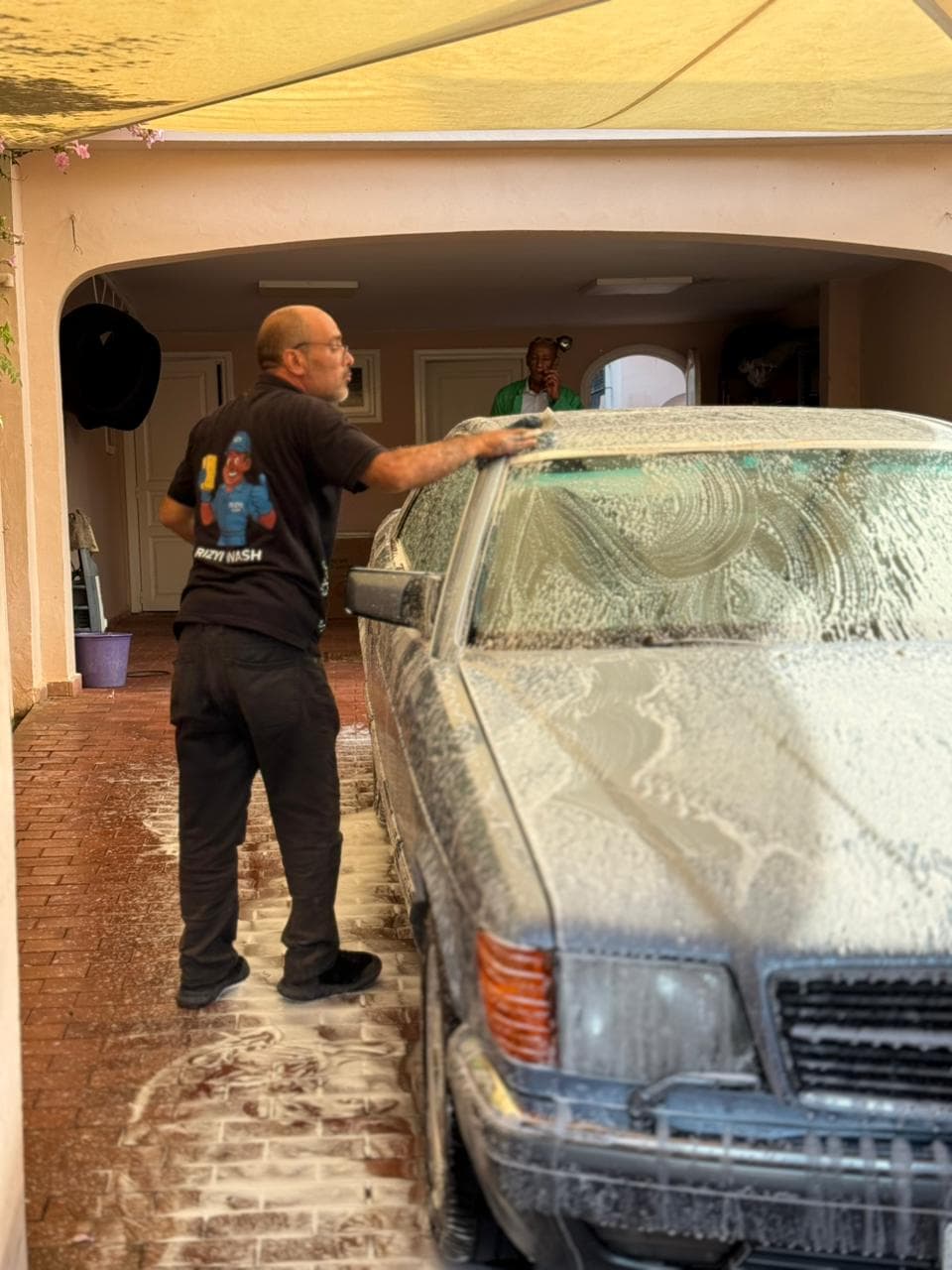 Man in black scrubs a vintage Mercedes Benz covered in soap suds in a driveway.