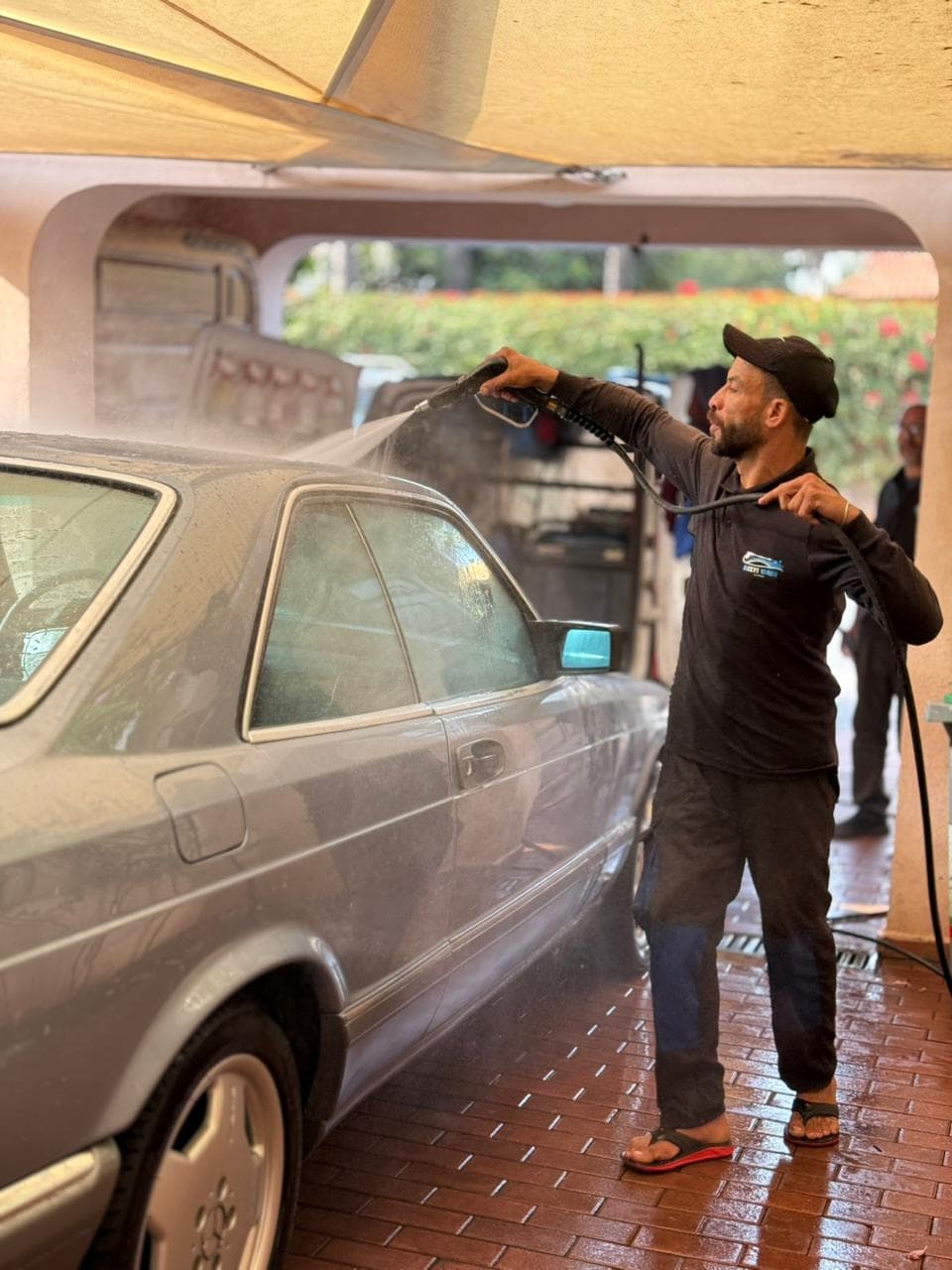 Man in black clothing pressure washing a silver classic car under a shaded driveway.