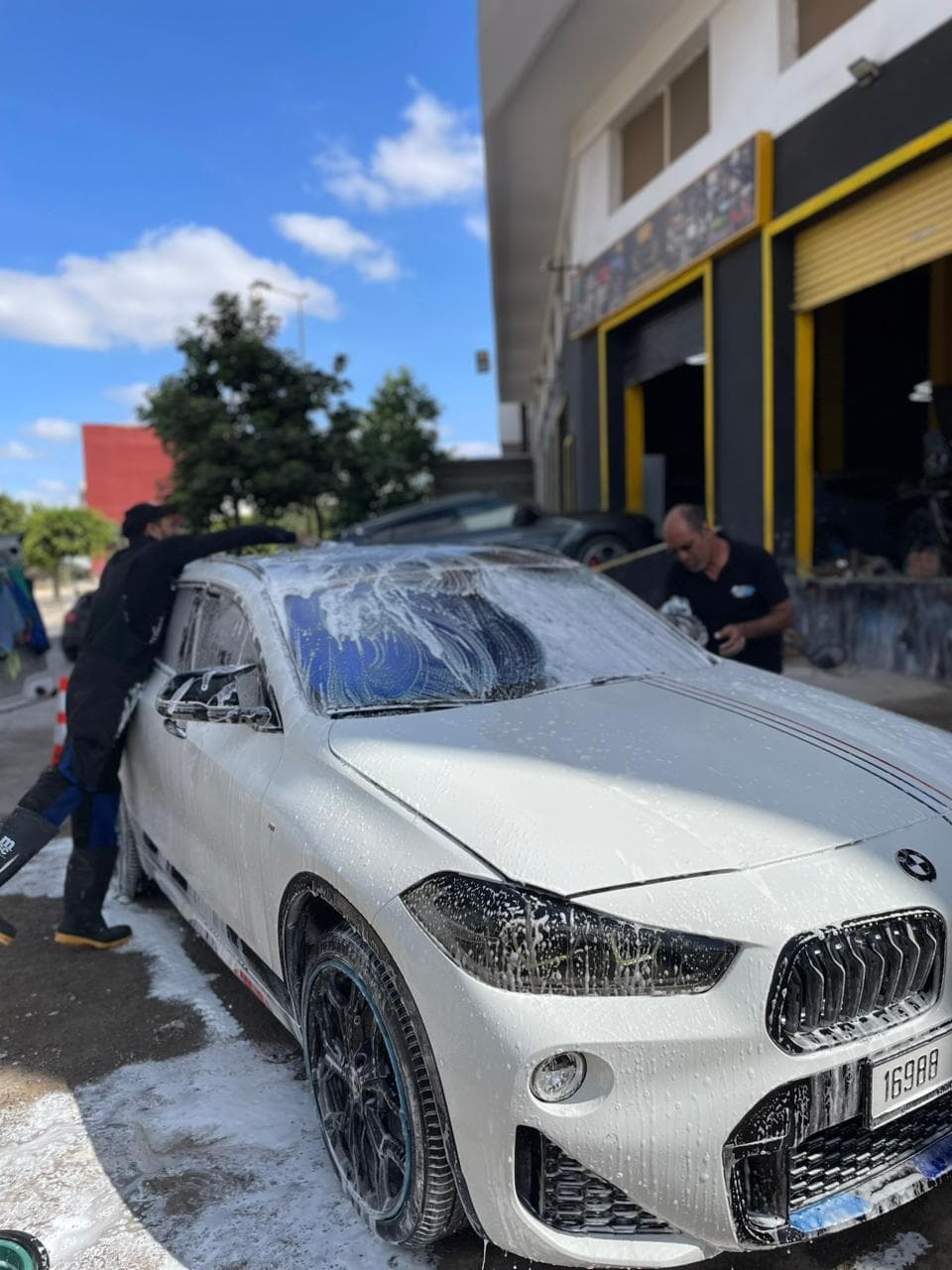 Two men washing a white BMW covered in soap foam at an outdoor car wash.