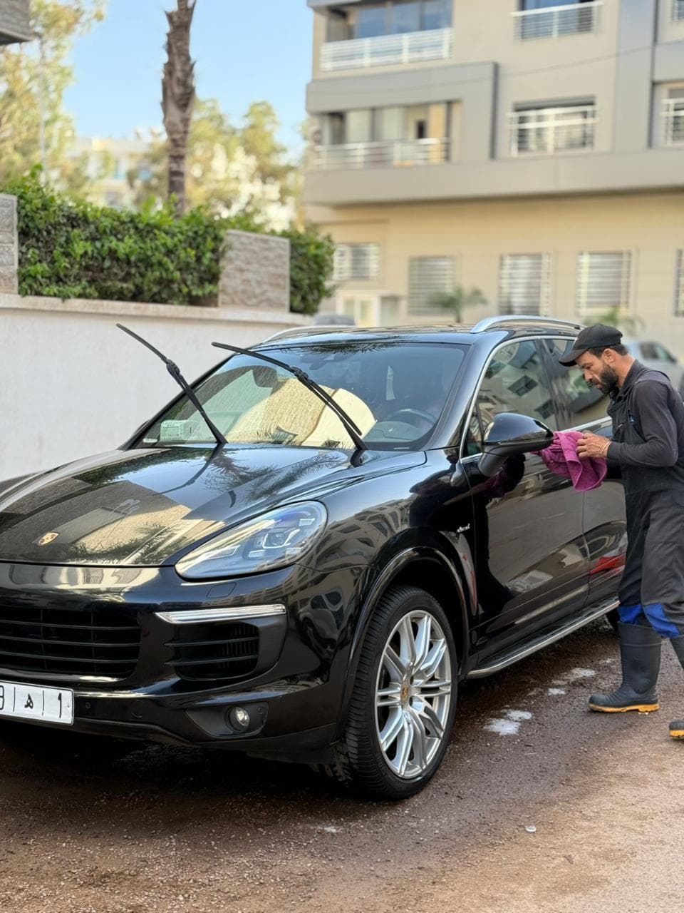 Man drying a black Porsche SUV with a pink cloth, windshield wipers raised, outdoors.