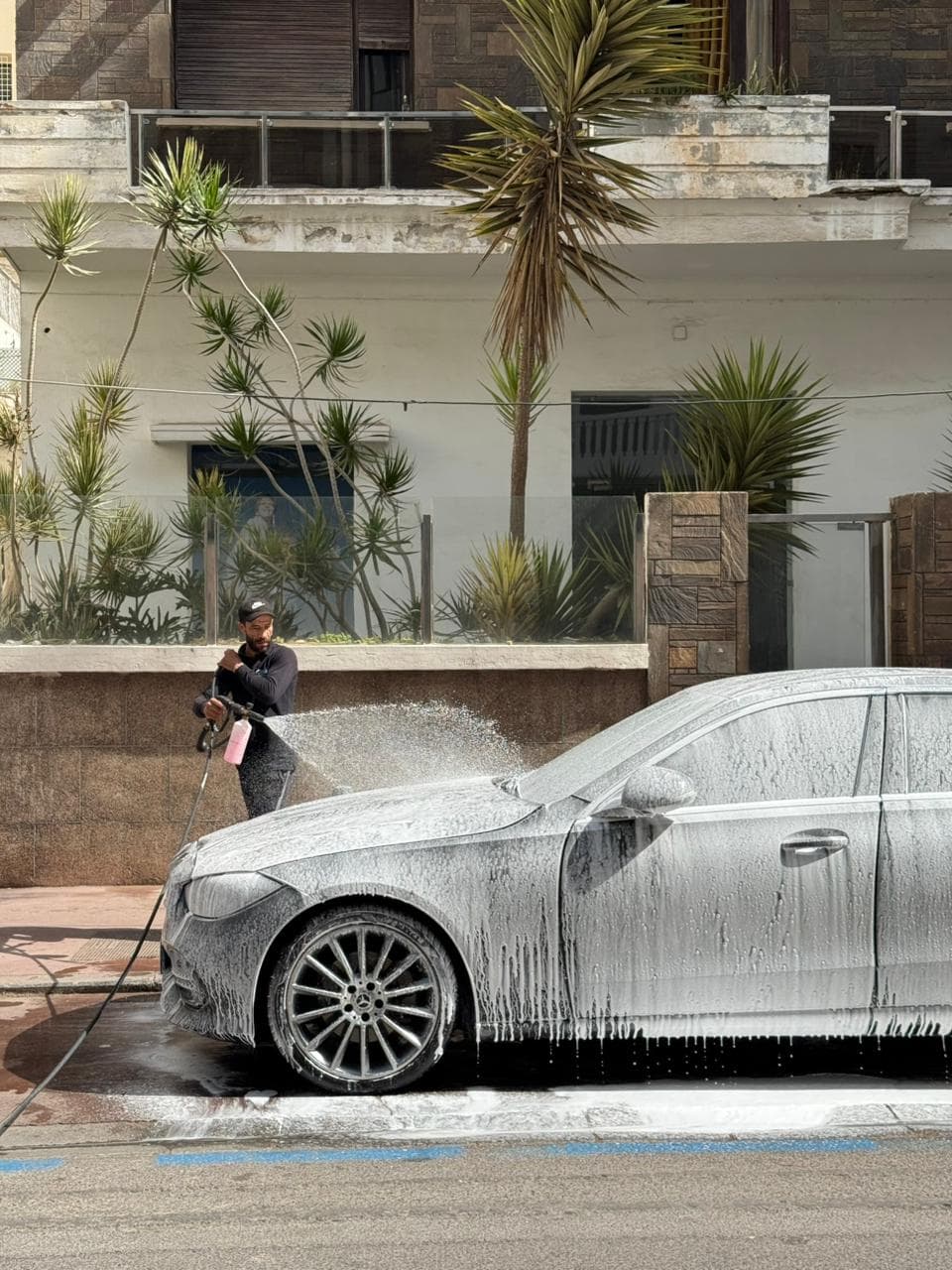 Man spraying thick white soap foam onto a silver car using a pressure washer.