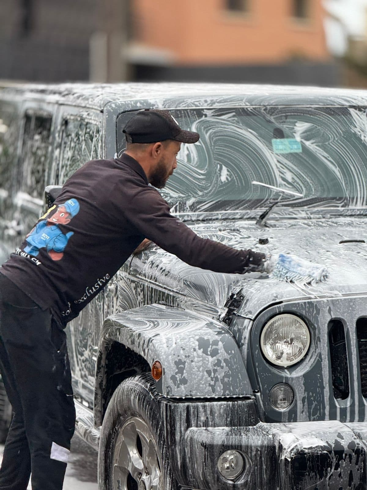Man in a black cap scrubbing a soapy grey Jeep Wrangler with a wash mitt.
