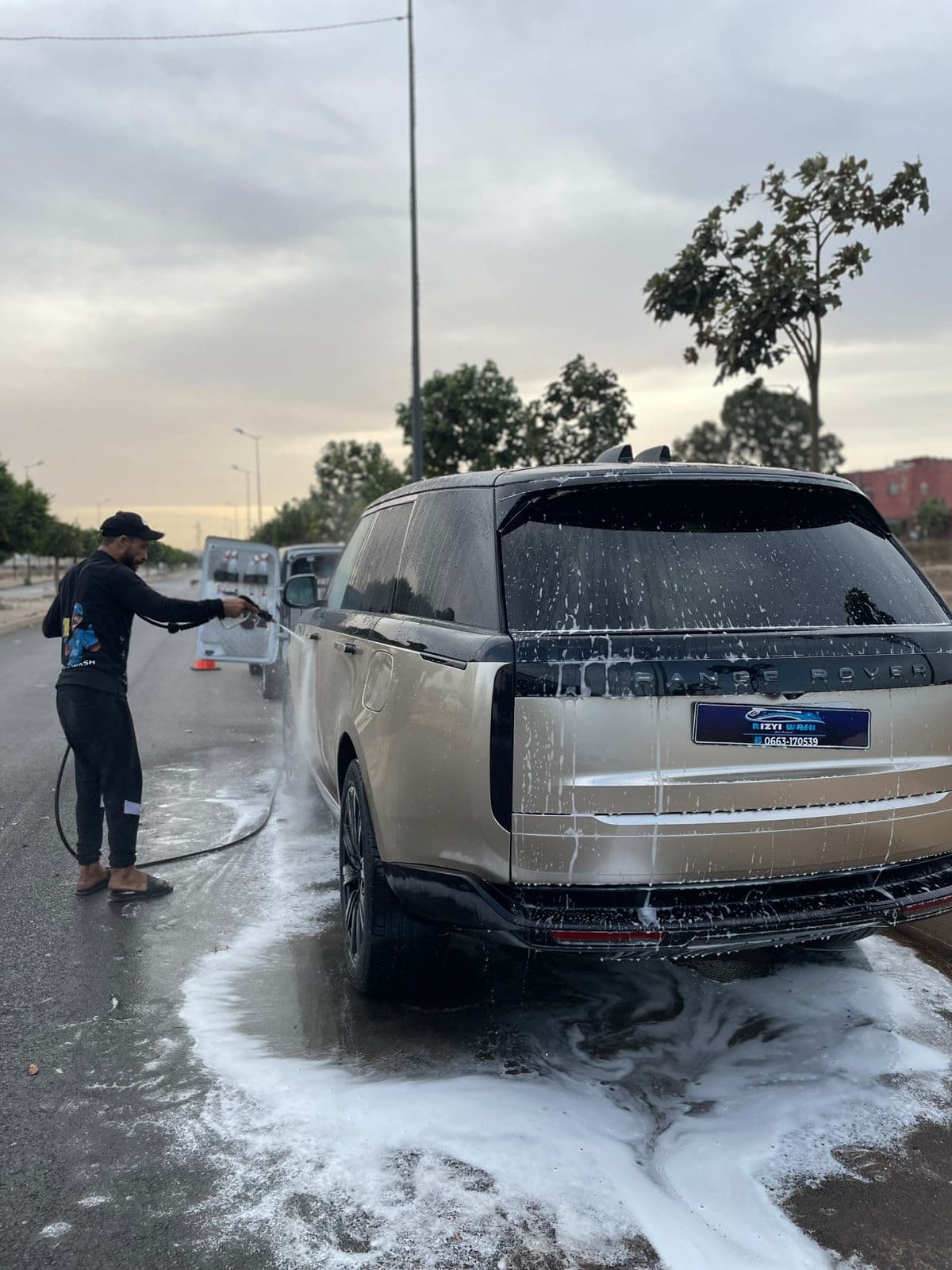 Man washes a soapy gold Range Rover with a pressure washer on a street.