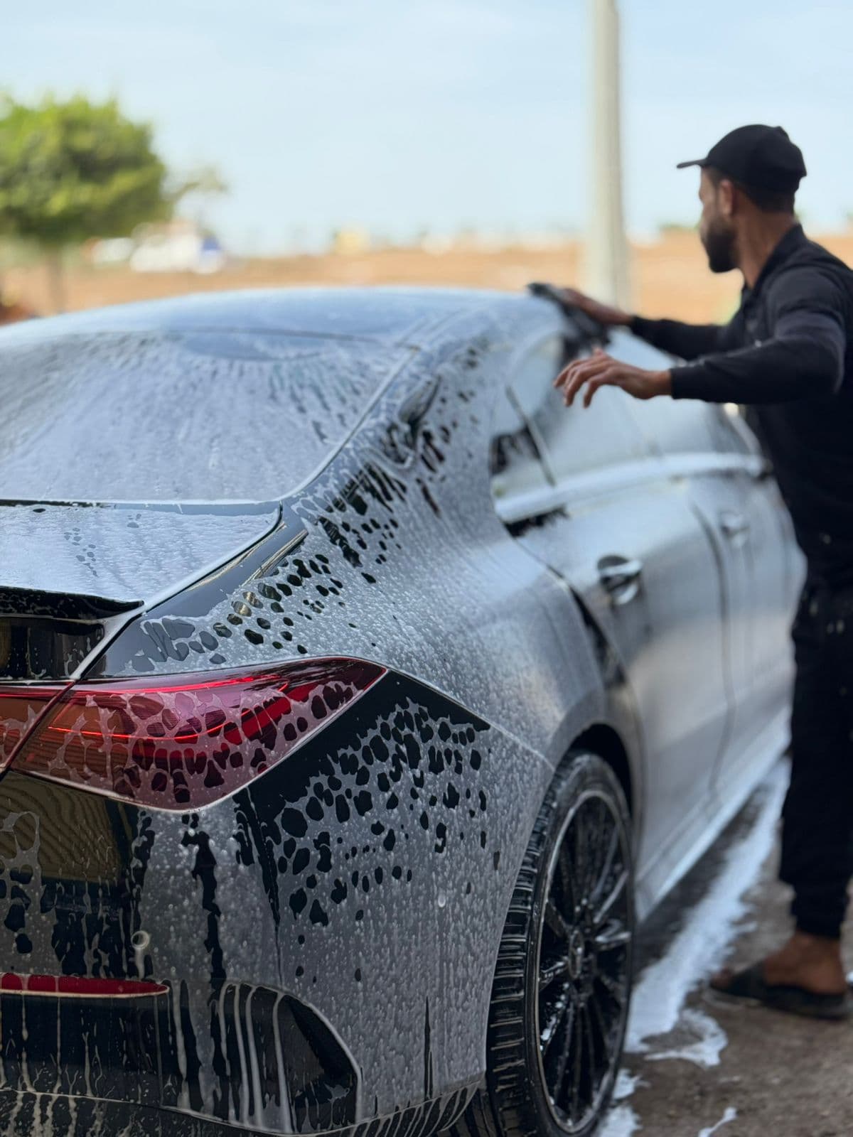Man in black clothing washing a black car covered in thick white soap suds.