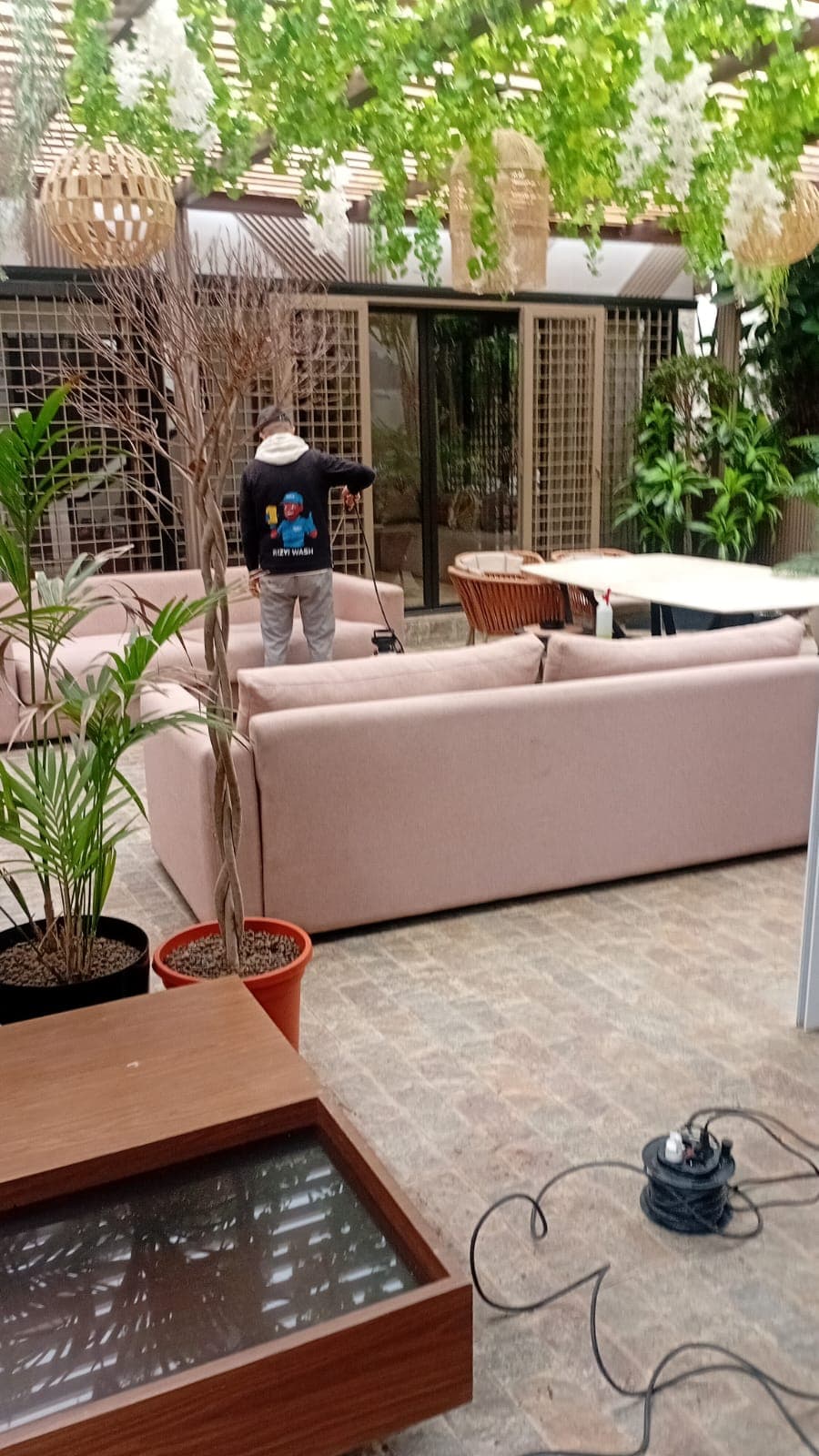 Professional cleaning a pink sofa in a stylish outdoor lounge area with hanging greenery.