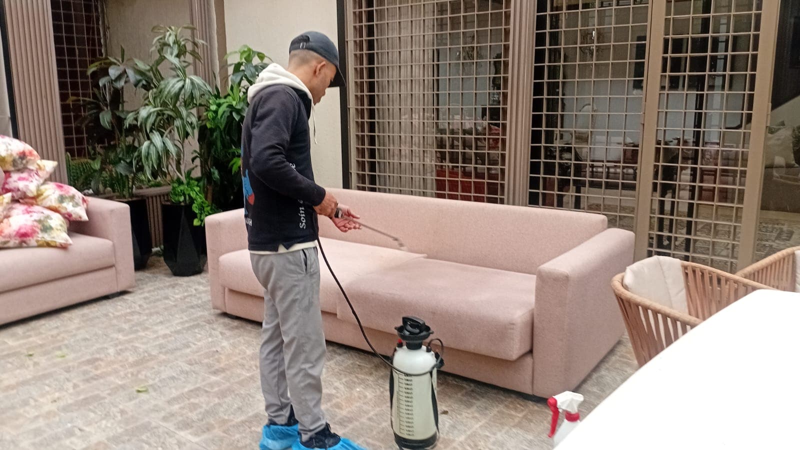 Man cleaning a light pink sofa with a handheld sprayer on a stone patio.
